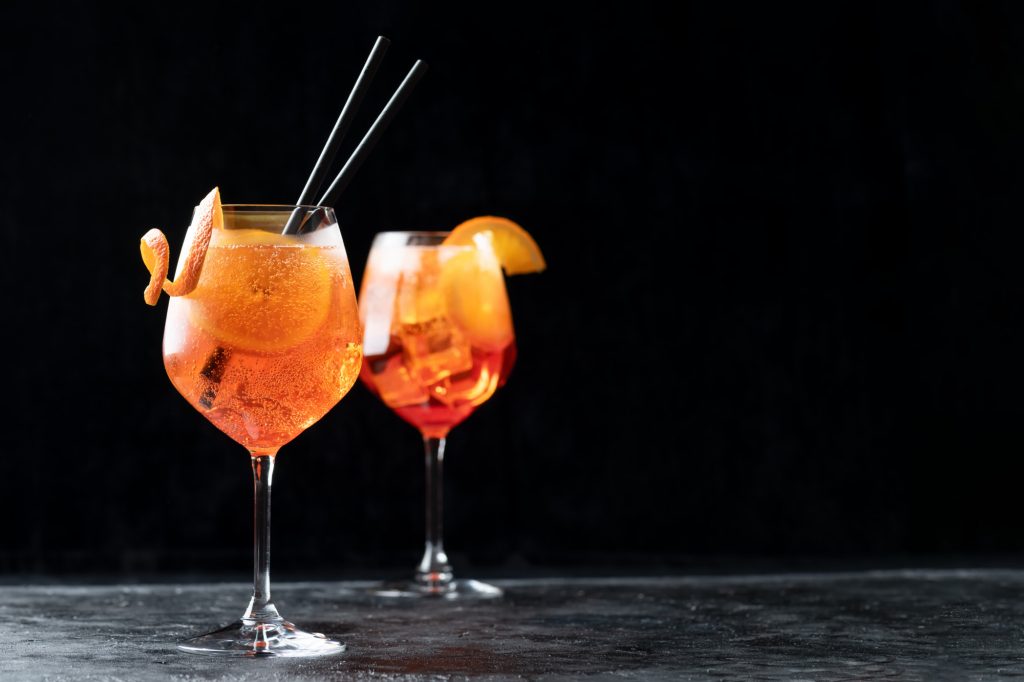 Un pahar de Aperol Spritz, cu gheață și garnisit cu o felie de portocală, plasat pe un fundal negru, evidențiat de o iluminare dramatică.