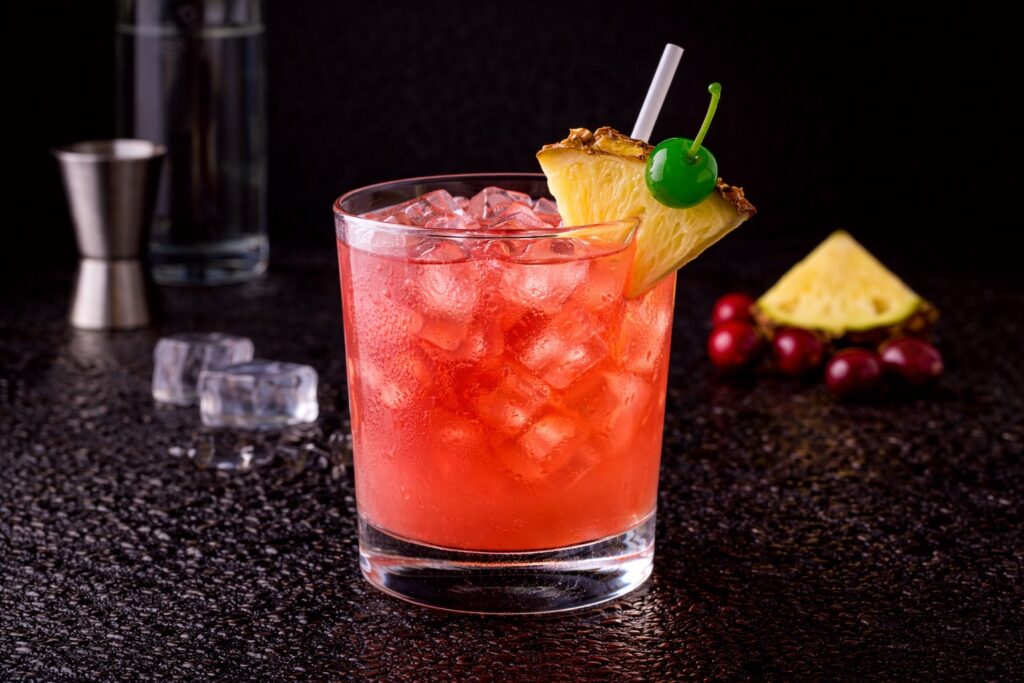 Cocktail Bay Breeze în pahar bucket cu gheață, vodcă, suc de merișoare și ananas, decorat cu felie de ananas pe fundal negru.