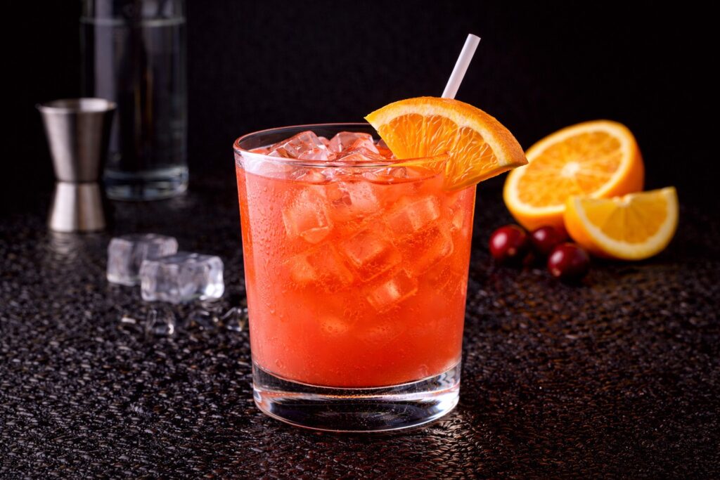 Cocktail Madras în pahar bucket cu gheață, vodcă, suc de portocale și suc de merișoare, decorat cu felie de portocală pe fundal negru.